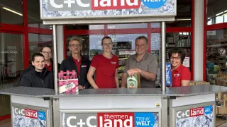 Five people stand behind a counter inside a store, smiling for the camera. They hold various products and one is holding a bottle of liquor. The sign above them reads C+C land and Trink Welt.