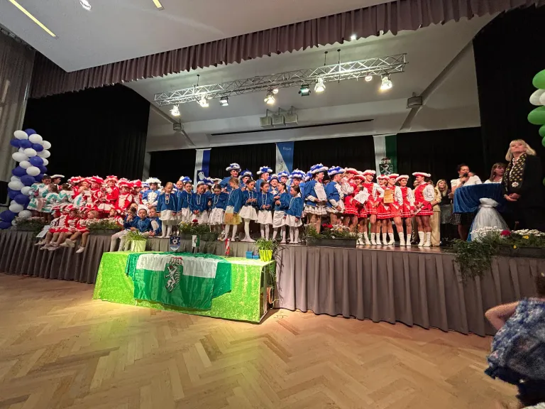 Eine Gruppe von Kindern in Kostümen steht auf der Bühne, einige tragen Hüte. Ein grüner Stoff mit einem Logo bedeckt einen Tisch davor. Scheinwerfer sind an der Decke befestigt.