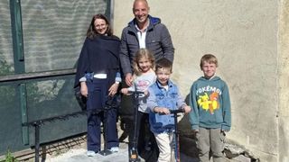 A family stands outside on a sidewalk. A woman in a shawl, a man in a jacket, and three children with scooters pose for a photo. The children are wearing sneakers.
