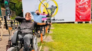 A man in a wheelchair aims a bow and arrow at a target during an archery event.
