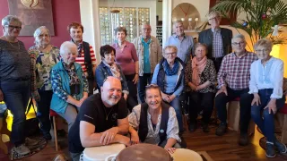 Eine Gruppe von Menschen ist drinnen versammelt. Einige sitzen, andere stehen hinter ihnen. Sie alle lächeln und scheinen für ein Gruppenfoto zu posieren. Ein Trommel ist im Vordergrund vorhanden.