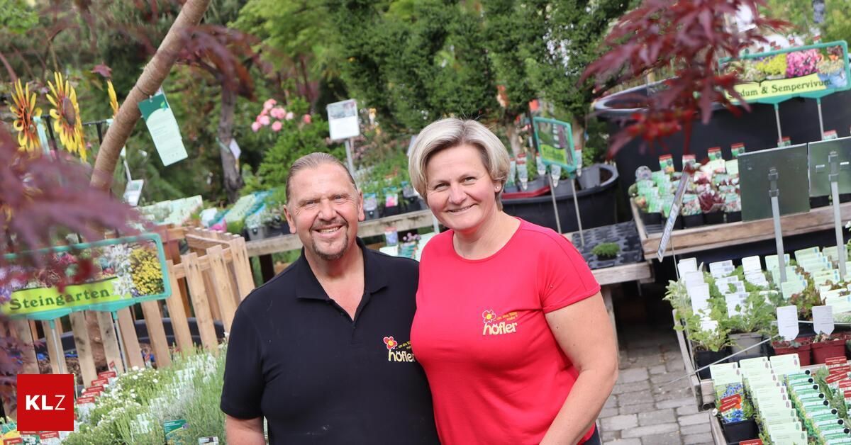 A man and a woman stand smiling in front of a garden, both wearing company polos. Behind them, various plants are displayed in trays.