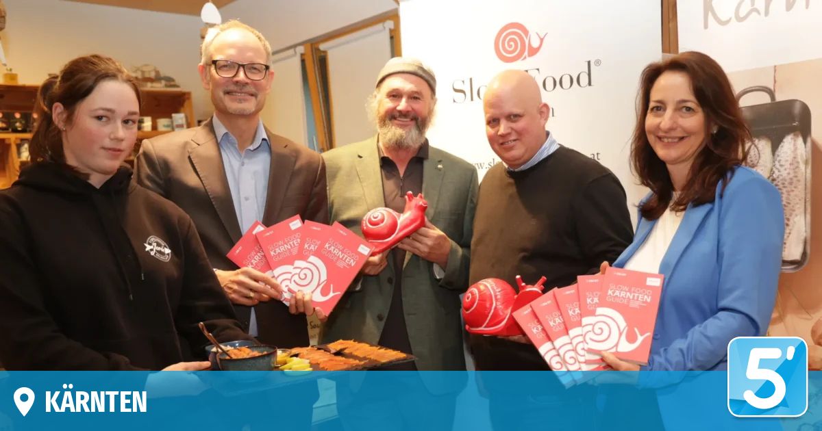 Fünf Personen posieren mit roten Slow-Food-Führern und Schneckenspielzeug. Sie stehen lächelnd im Innenbereich. Vor ihnen steht ein Tisch mit Essen und ein blauer Schriftzug dahinter, der 'Slow Food' lautet.