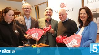 Fünf Personen posieren mit roten Slow-Food-Führern und Schneckenspielzeug. Sie stehen lächelnd im Innenbereich. Vor ihnen steht ein Tisch mit Essen und ein blauer Schriftzug dahinter, der 'Slow Food' lautet.