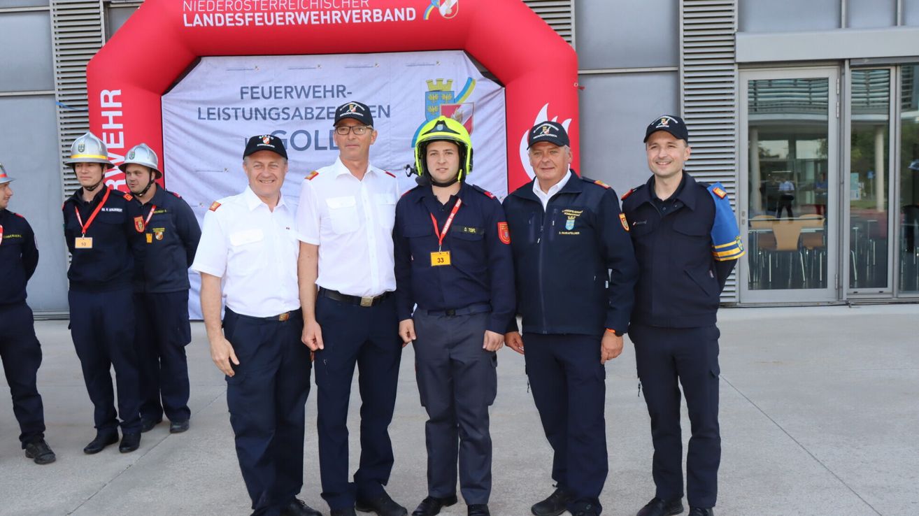 Fünf Feuerwehrleute stehen zusammen vor einem Banner. Einer trägt einen Helm, ein anderer hat eine Medaille. Sie sind alle in Uniform.