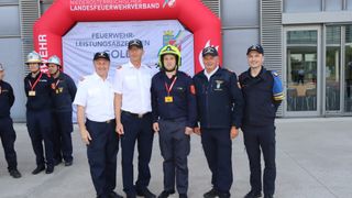 Fünf Feuerwehrleute stehen zusammen vor einem Banner. Einer trägt einen Helm, ein anderer hat eine Medaille. Sie sind alle in Uniform.