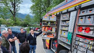 Eine Gruppe von Menschen, einschließlich eines Mannes, der zeigt, beobachtet ein Feuerwehrauto mit einem offenen Staufach voller Werkzeuge und Ausrüstung.