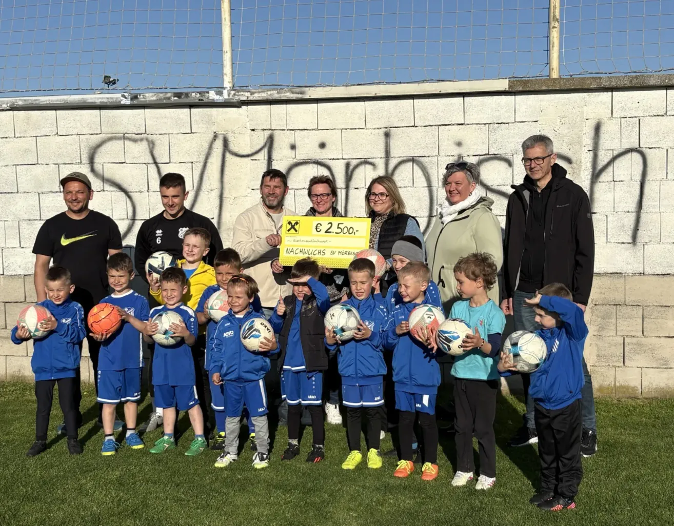 Eine Gruppe junger Jungen in blauen Uniformen und ihren Trainern posiert mit Fußballbällen und einem großen Scheck über 2.500 Euro vor einer Wand.
