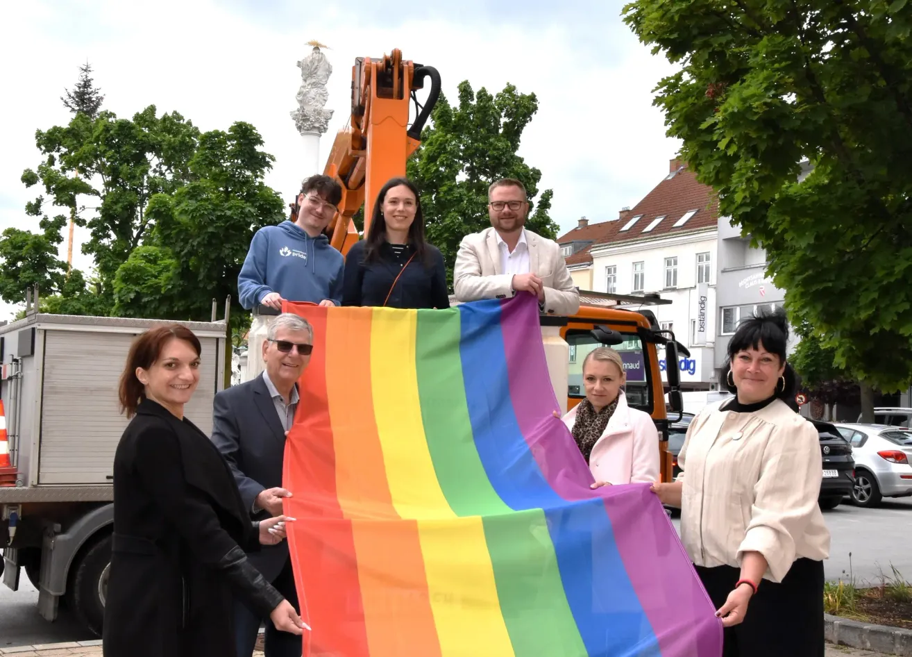 Eine Gruppe von Menschen, die eine große Regenbogenflagge vor einem Baufahrzeug halten, steht an einem sonnigen Tag in einer städtischen Umgebung.