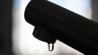 Ein einzelner Wassertropfen hängt von einem schwarzen Wasserhahn, beleuchtet von einem sanften Licht.