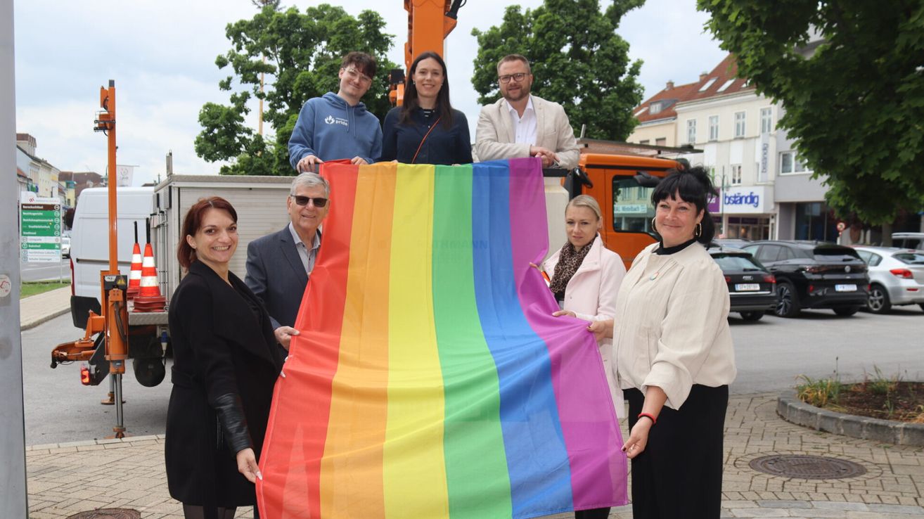 Eine Gruppe von Menschen hält eine große Regenbogenflagge im Freien. Eine Person steht hinter einem Kran.