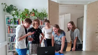 Five women in a room, with one woman using a computer. The woman in the middle is smiling, while the others are looking at the computer.
