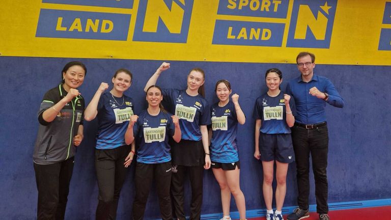 Fünf Frauen in identischen blauen Shirts stehen vor einem Banner, lächeln und heben ihre Fäuste. Eine hat eine Medaille um den Hals. Hinter ihnen steht ein Mann mit Brille und Gürtel.