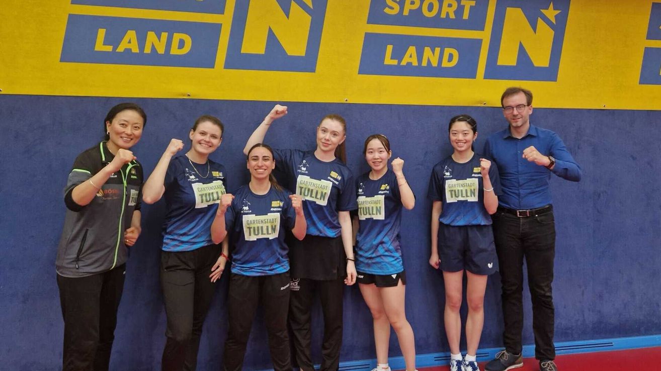Fünf Frauen in identischen blauen Shirts stehen vor einem Banner, lächeln und heben ihre Fäuste. Eine hat eine Medaille um den Hals. Hinter ihnen steht ein Mann mit Brille und Gürtel.