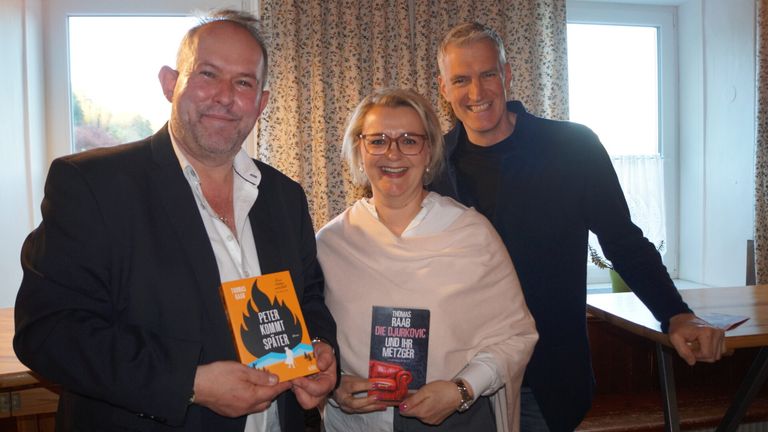 Three people are smiling for a photo in a room. Two of them are holding books, and one is wearing a watch. The book covers are visible, one is titled 'Peter kommt spater' and the other 'Die Djurkovic und ihr Metzger'.
