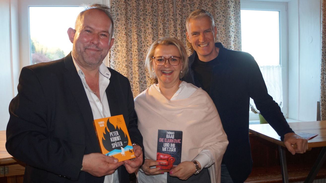 Three people are smiling for a photo in a room. Two of them are holding books, and one is wearing a watch. The book covers are visible, one is titled 'Peter kommt spater' and the other 'Die Djurkovic und ihr Metzger'.