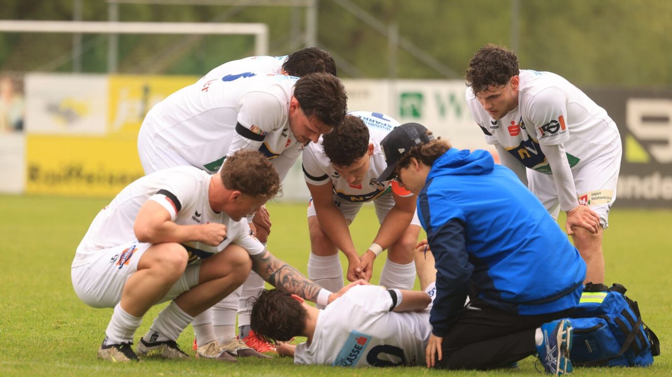 Eine Fußballmannschaft in weißen Trikots ist um einen verletzten Spieler auf einem Rasenfeld versammelt, mit einem Tor und Zaun im Hintergrund.
