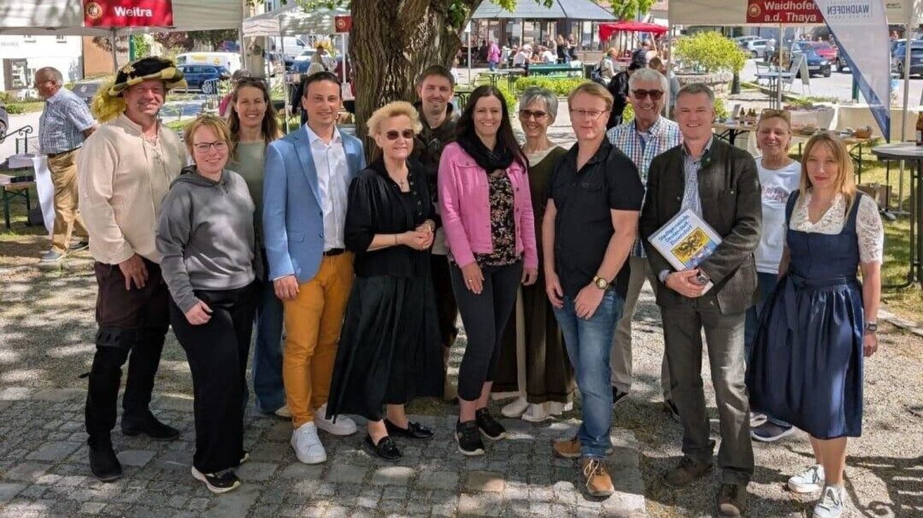 Eine Gruppe von Menschen, sowohl Männer als auch Frauen, stehen vor einem Baum, lächeln und schauen in die Kamera. Im Hintergrund sind Zelte und Menschen zu sehen.