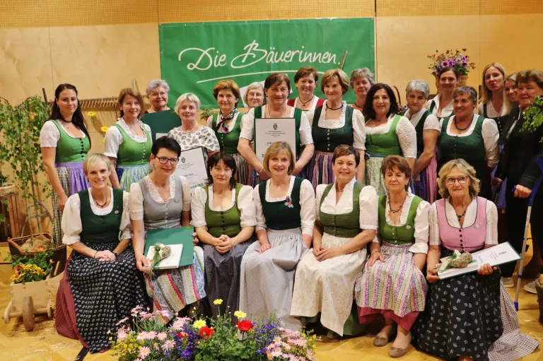 A group of women wearing traditional green and white clothing are posing for a photo with certificates and flowers in front of a green banner.