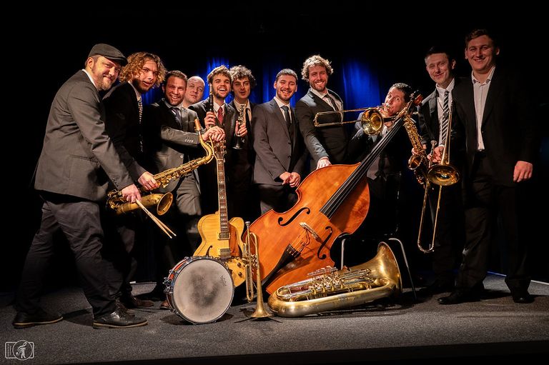 Eine Gruppe von Musikern posiert auf der Bühne mit verschiedenen Instrumenten, darunter Trompeten, Saxophone und eine Bass. Sie sind alle in formellen Anzügen gekleidet.