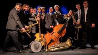 Eine Gruppe von Musikern posiert auf der Bühne mit verschiedenen Instrumenten, darunter Trompeten, Saxophone und eine Bass. Sie sind alle in formellen Anzügen gekleidet.