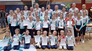 Eine Gruppe junger Turnerinnen hält Urkunden und Medaillen in den Händen und trägt passende blaue und weiße Uniformen. Sie posieren für ein Foto in einer Turnhalle.