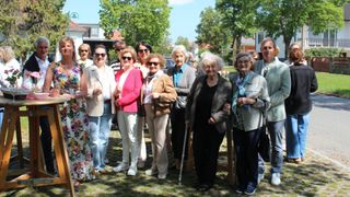 Eine Gruppe älterer Menschen, alle mit Sonnenbrillen, stehen auf einem grünen Rasen, wahrscheinlich in einem Park. Sie lächeln alle und scheinen für ein Foto zu posieren.