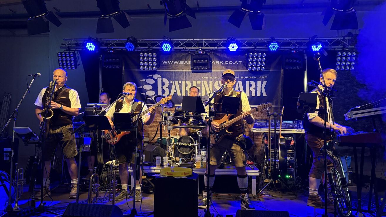 A band performs on a stage with blue lights. Two men play guitars, and a man plays drums. A banner behind them reads 'BARRENSARK'.