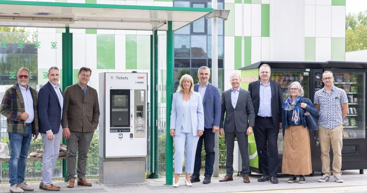 Six people stand in front of a ticket machine at a bus stop. Behind them is a building with a green and white pattern.