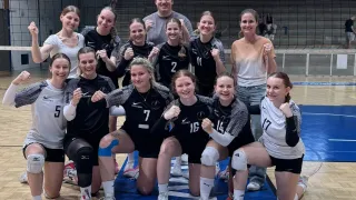 Ein Volleyballteam feiert auf dem Spielfeld, trägt schwarze und weiße Trikots, mit den Nummern 2, 5, 7, 14, 16 und 17 sichtbar. Dahinter befindet sich ein Volleyballnetz und leere Tribünen.