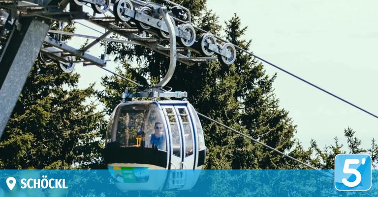 Eine Gondelbahn mit zwei Personen ist an Seilen von einer Metallstruktur aufgehängt und fährt durch einen Wald.