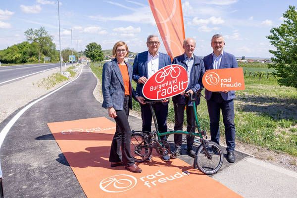 Four people stand next to a new cycling path in a scenic area with trees and fields. They hold signs that say 'Burgenland radelt' and 'rad freude'.