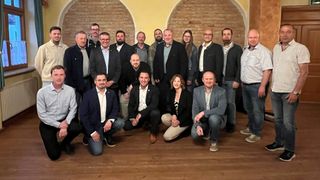 Eine Gruppe von Menschen in formeller Kleidung posiert für ein Foto vor einer Ziegelwand. Einige sitzen, andere stehen.