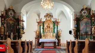 Der Innenraum einer Kirche mit einem großen, verzierten Altar, Statuen und einer Kristallleuchte. Bänke sind angeordnet und ein roter Teppich führt zum Altar.
