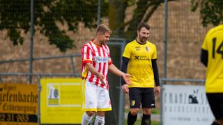 Zwei männliche Fußballspieler gehen und unterhalten sich auf einem Feld. Beide tragen Trikots mit Werbung. Der Spieler auf der linken Seite trägt ein rot-weißes Trikot. Der Spieler auf der rechten Seite trägt ein gelbes Trikot. Hinter ihnen befindet sich ein Metallzaun mit einem gelben Banner.
