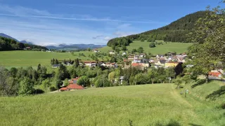 Ein malerisches Dorf in einem Tal, umgeben von üppigen grünen Hügeln und Bergen, unter einem klaren blauen Himmel.