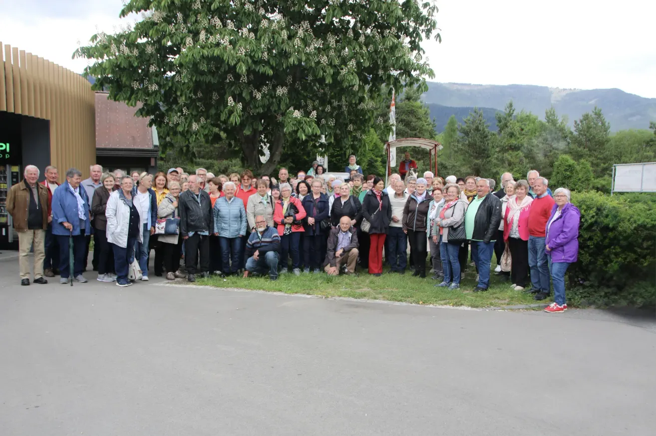 Eine große Gruppe von Menschen, überwiegend ältere, posiert für ein Foto in einem Park mit einem großen Baum und Bergen im Hintergrund.