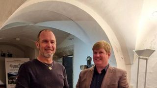 Zwei Männer stehen Seite an Seite unter einem Bogen und lächeln. Ein Mann trägt eine Halskette und ein dunkles Shirt, der andere einen hellen Anzug.