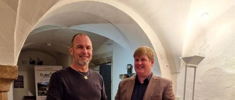 Zwei Männer stehen Seite an Seite unter einem Bogen und lächeln. Ein Mann trägt eine Halskette und ein dunkles Shirt, der andere einen hellen Anzug.