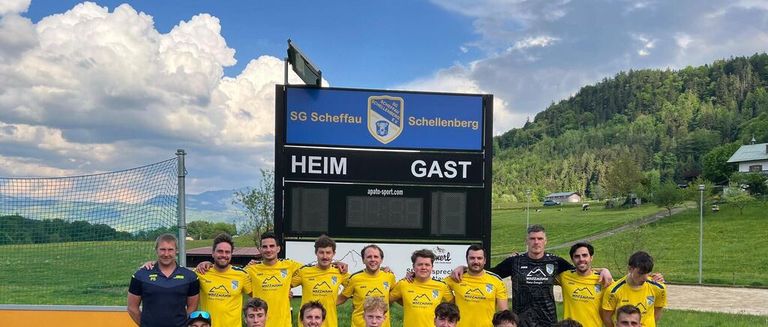 Eine Fußballmannschaft posiert vor einer Anzeigetafel mit 'HEIIM' und 'GAST'. Das Team trägt gelbe Trikots. Der Himmel ist blau mit Wolken.