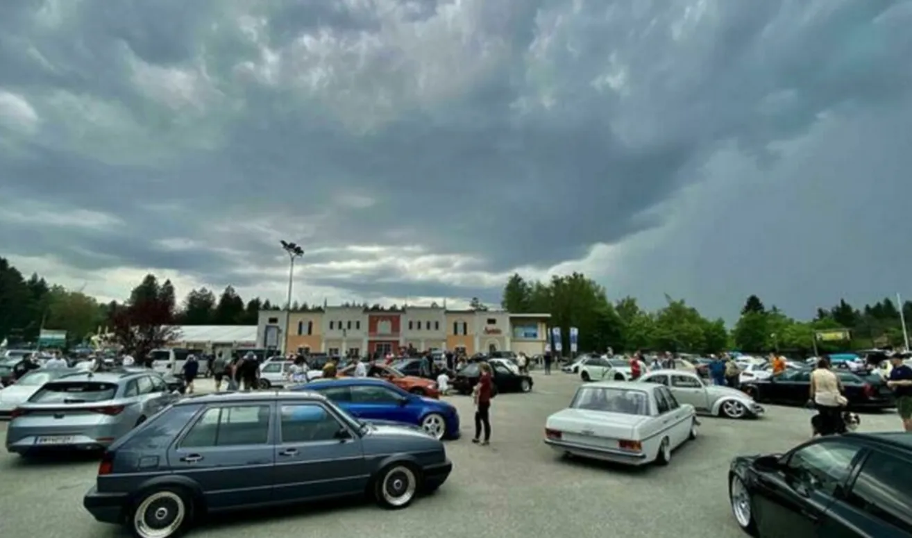 Ein Parkplatz voller Autos. Menschen gehen und stehen herum. Im Hintergrund sind Gebäude. Der Himmel ist bewölkt.