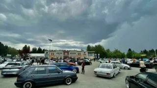Ein Parkplatz voller Autos. Menschen gehen und stehen herum. Im Hintergrund sind Gebäude. Der Himmel ist bewölkt.