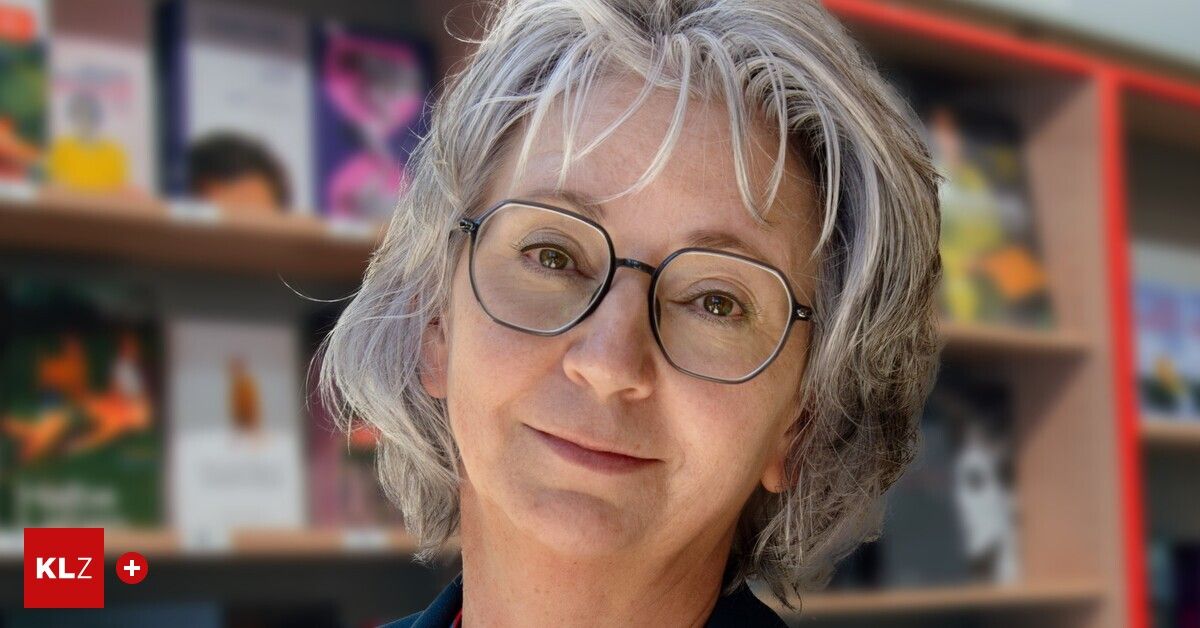 Eine Frau mit grauen Haaren und einer Brille lächelt und steht vor einem Bücherregal.