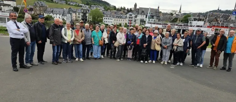 Eine Gruppe älterer Erwachsener steht vor einem Gebäude und posiert lächelnd für ein Foto. Dahinter befinden sich Gebäude mit Giebeldächern.