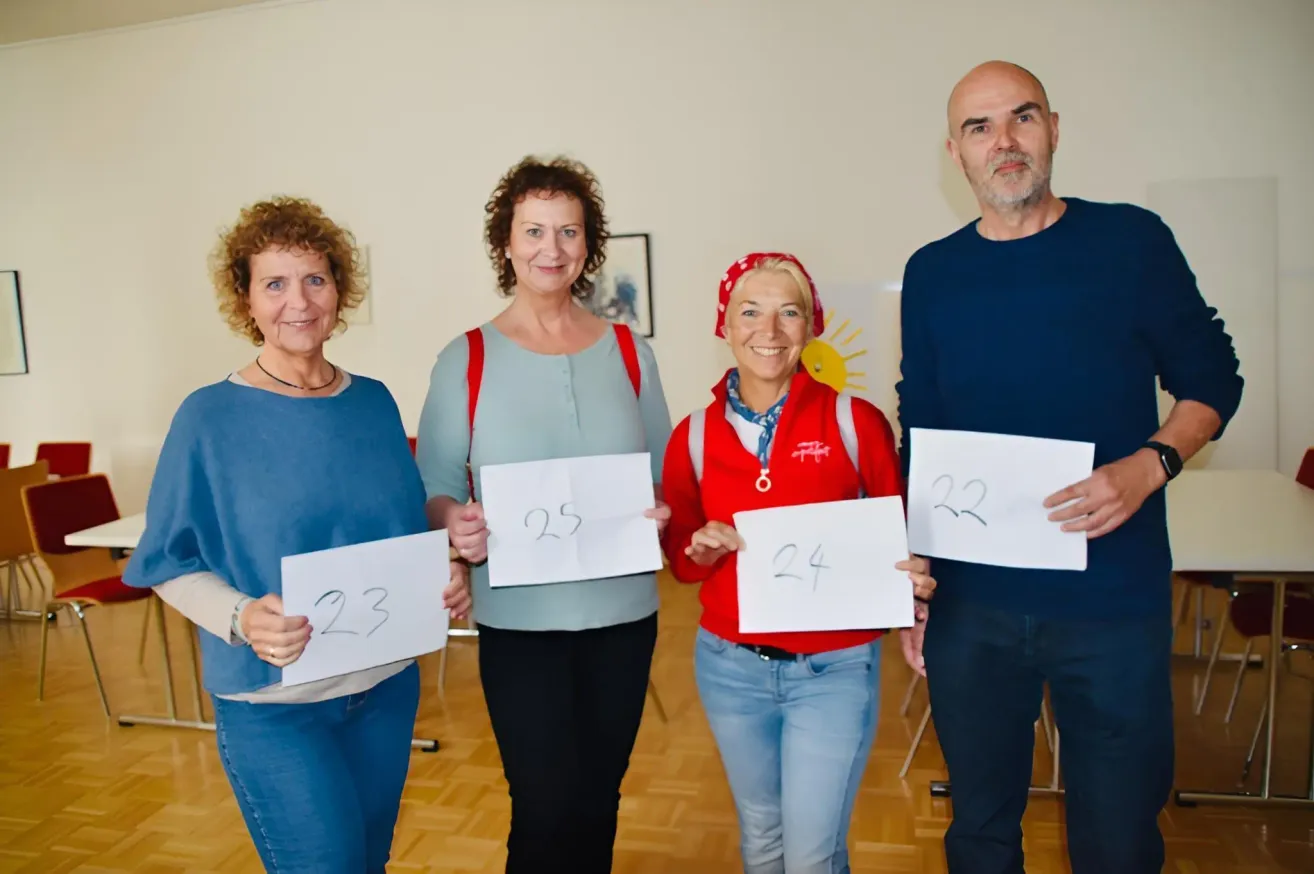 Vier Erwachsene, zwei Frauen und zwei Männer, stehen zusammen und halten weiße Schilder mit Nummern. Die Frau auf der linken Seite trägt ein blaues Shirt und Jeans, während der Mann auf der rechten Seite ein dunkelblaues Shirt trägt.