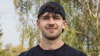 Ein Mann lächelt und posiert für ein Foto, trägt einen schwarzen Hut und ein schwarzes Shirt. Hinter ihm sind Bäume.