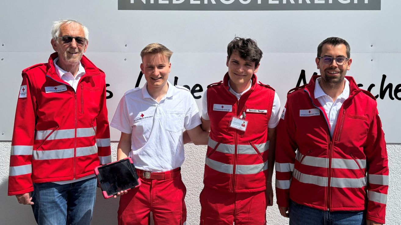 Vier Personen, gekleidet in roten und weißen Uniformen, stehen zusammen vor einer weißen Wand. Der Mann auf der linken Seite hält ein Tablet, während der Mann neben ihm eine rote Jacke mit weißen Streifen trägt.