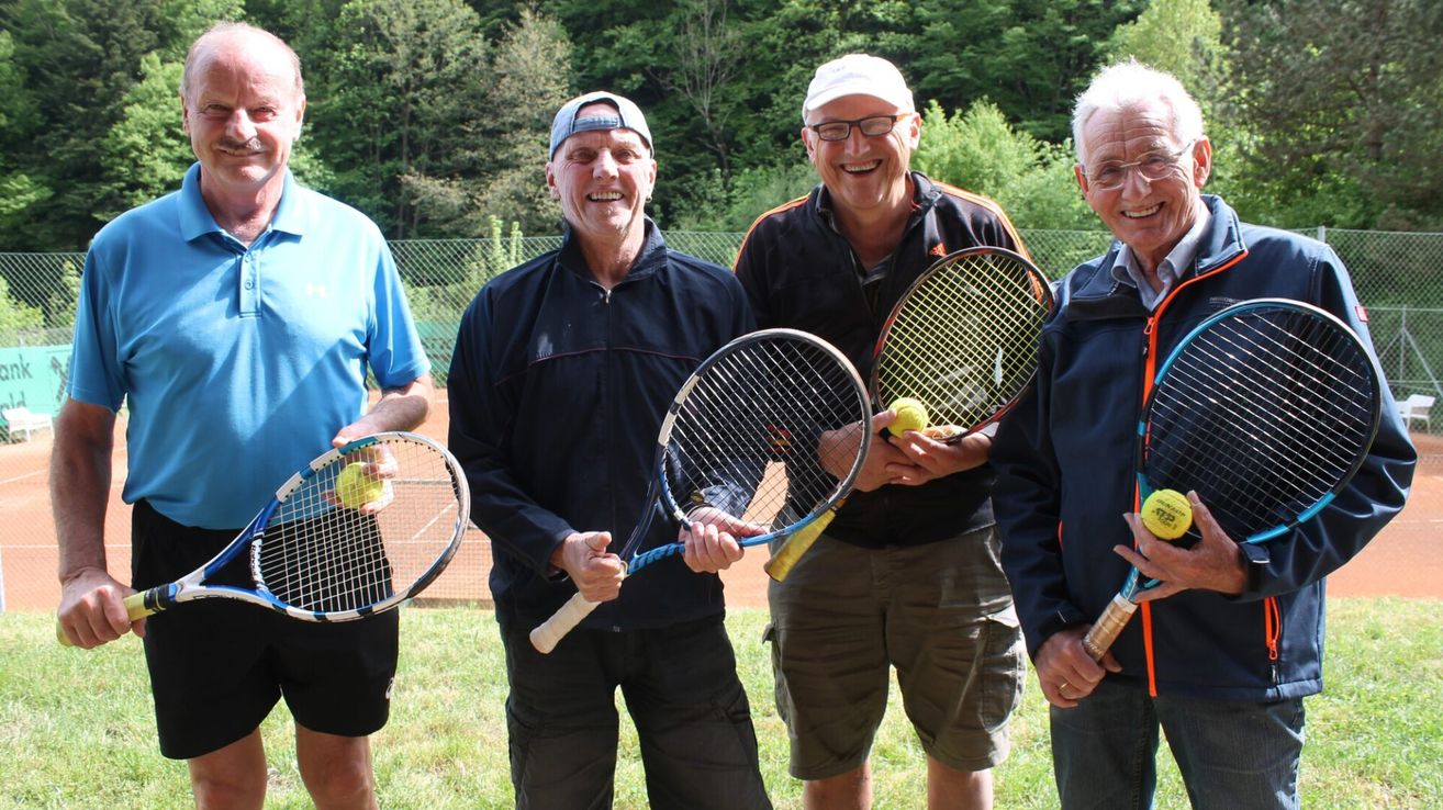 Vier Männer stehen auf einem Tennisplatz mit Schlägern und halten Tennisbälle in den Händen. Sie tragen Freizeitkleidung und lächeln, was darauf hindeutet, dass sie ihre Zeit gemeinsam genießen.