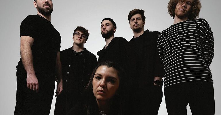 Eine Gruppe von Bandmitgliedern in schwarzer und weißer Kleidung steht für ein Foto zusammen. Die Frau in der Mitte schaut nach vorne, während die anderen zur Seite schauen.