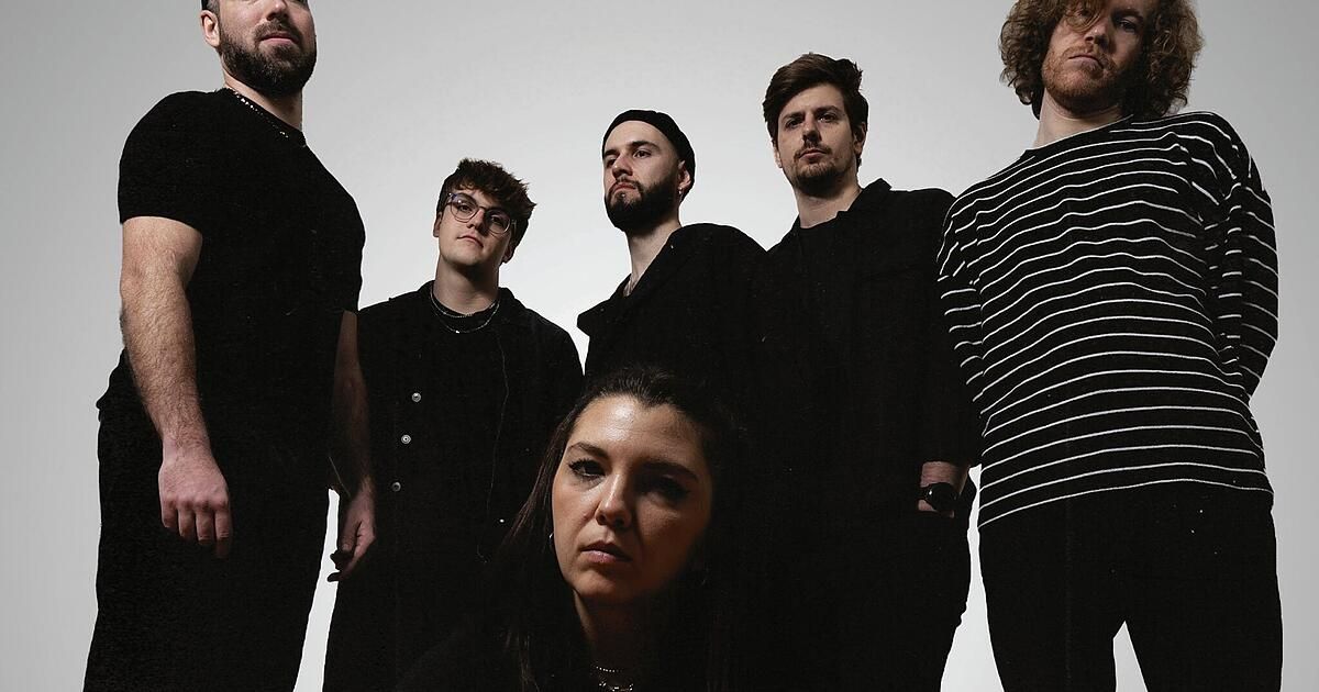 Eine Gruppe von Bandmitgliedern in schwarzer und weißer Kleidung steht für ein Foto zusammen. Die Frau in der Mitte schaut nach vorne, während die anderen zur Seite schauen.